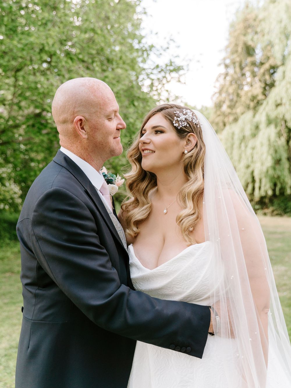 A bride in a flowing white dress and veil shares an intimate moment with groom in black suit outdoors among willow trees.