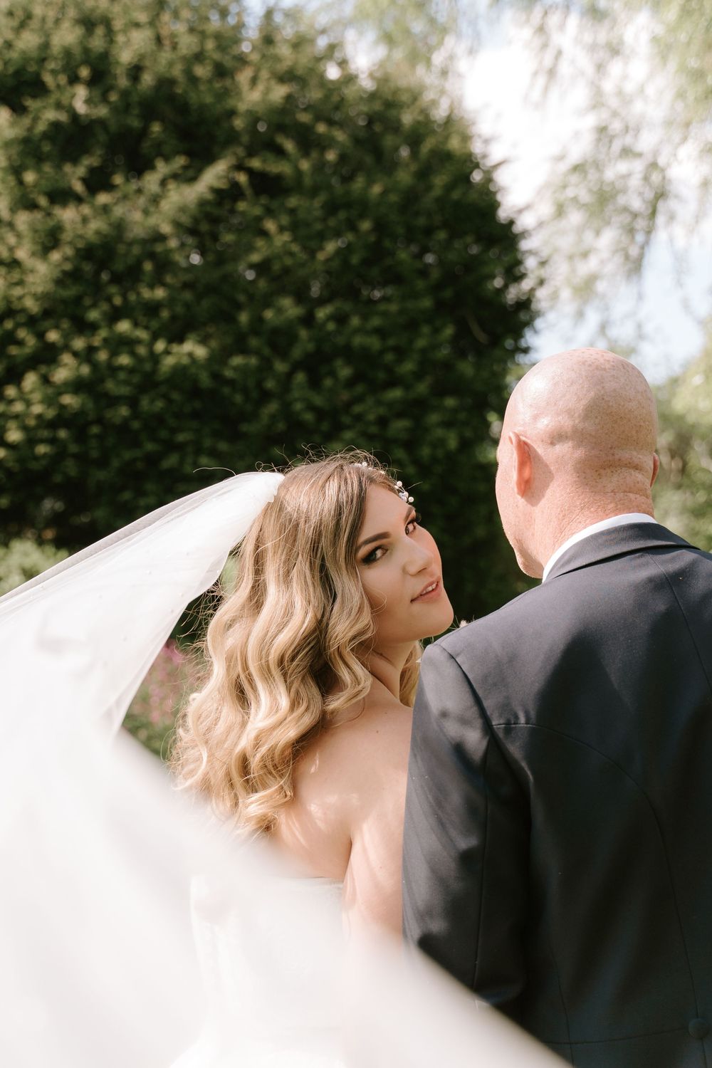 A beautiful bride in a white gown and flowing veil stands with a groom in black suit against a soft outdoor backdrop.