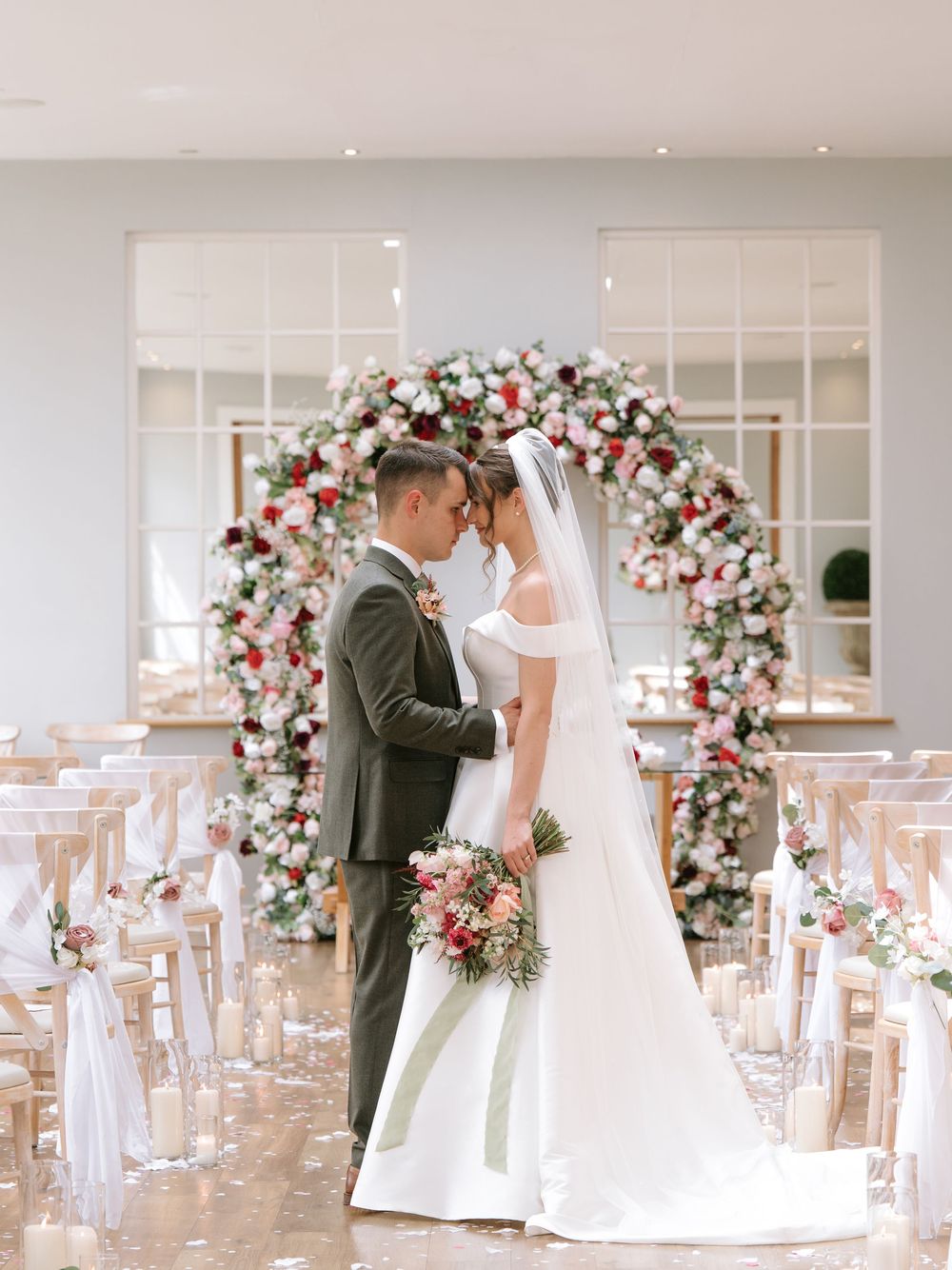 A romantic wedding ceremony setup with a floral arch featuring pink and white roses, surrounded by elegant ghost chairs.