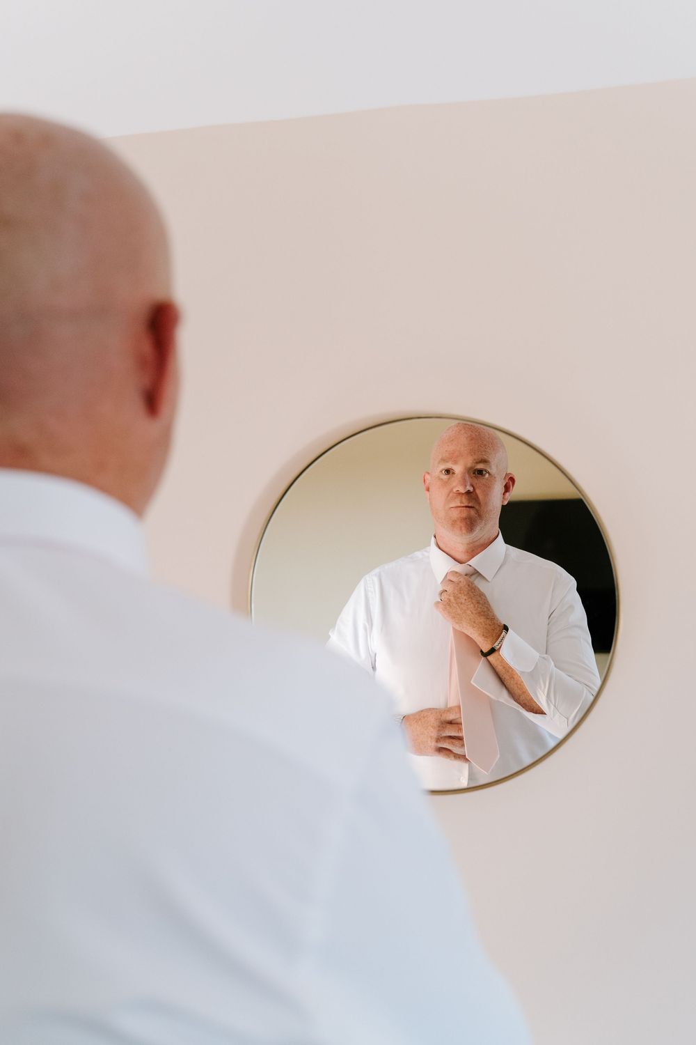A reflection in a round mirror shows someone adjusting a white dress shirt and tie.