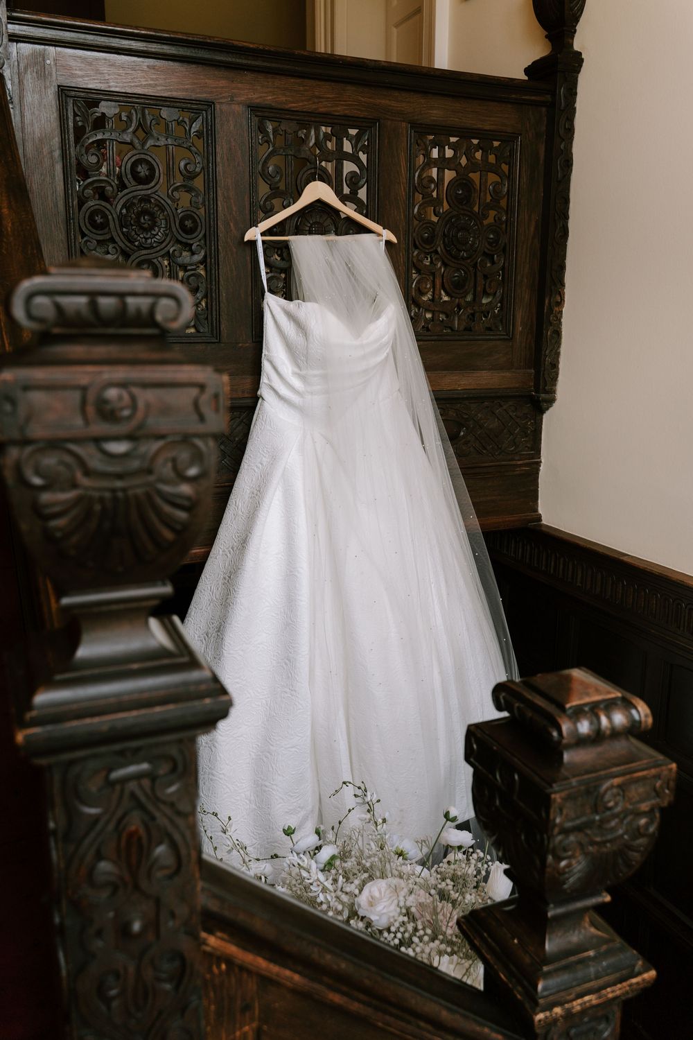 An elegant white wedding gown with delicate embroidery hangs on a wooden hanger against ornate vintage carved wooden railings.