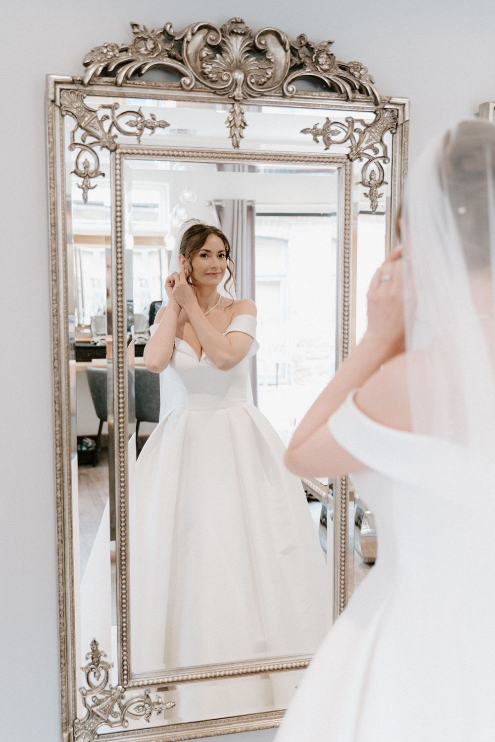 A reflection in an ornate silver baroque mirror shows a person in an elegant white wedding dress adjusting their veil.