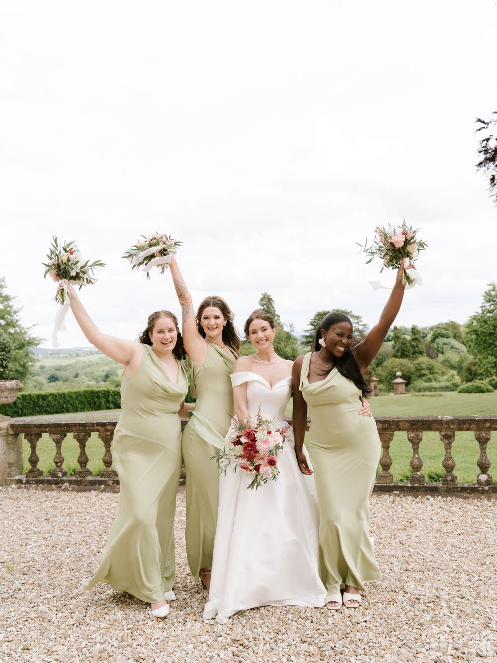Wedding party in matching sage green dresses poses joyfully holding bouquets on outdoor stone terrace.