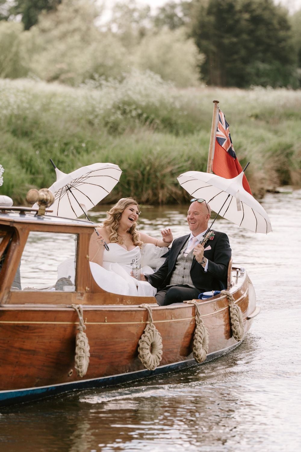 A couple in a wooden boat on a river hold white parasols beside a British maritime flag on a sunny day.