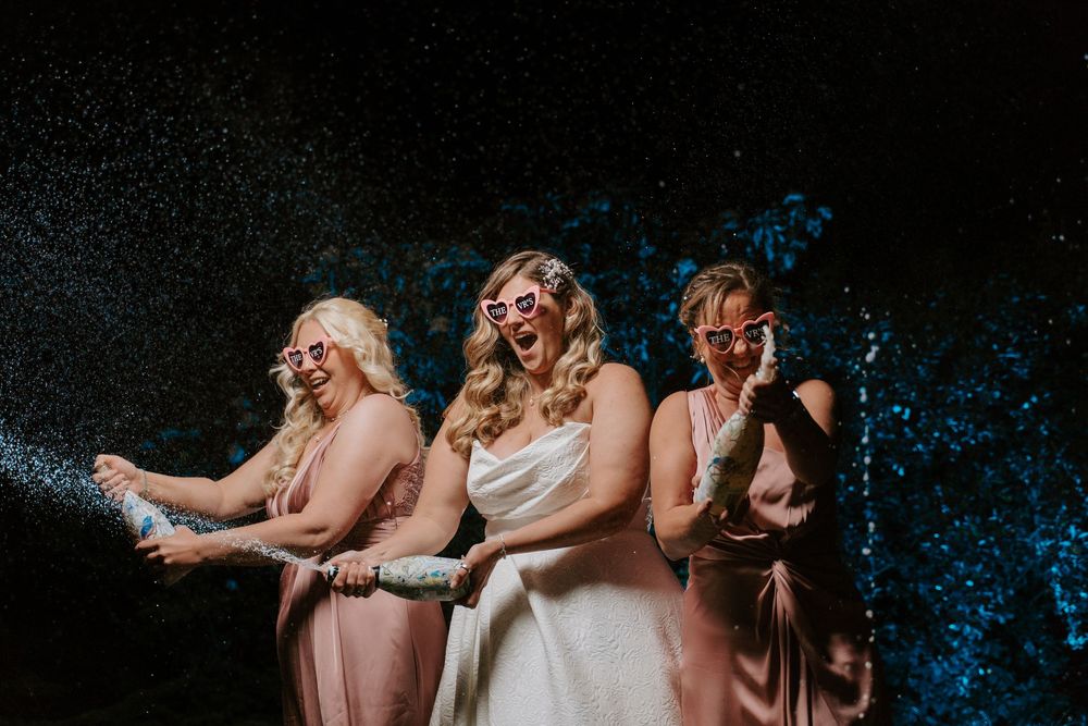 Three people in pink and white outfits react with joy and laughter while being showered with glitter against a dark background.