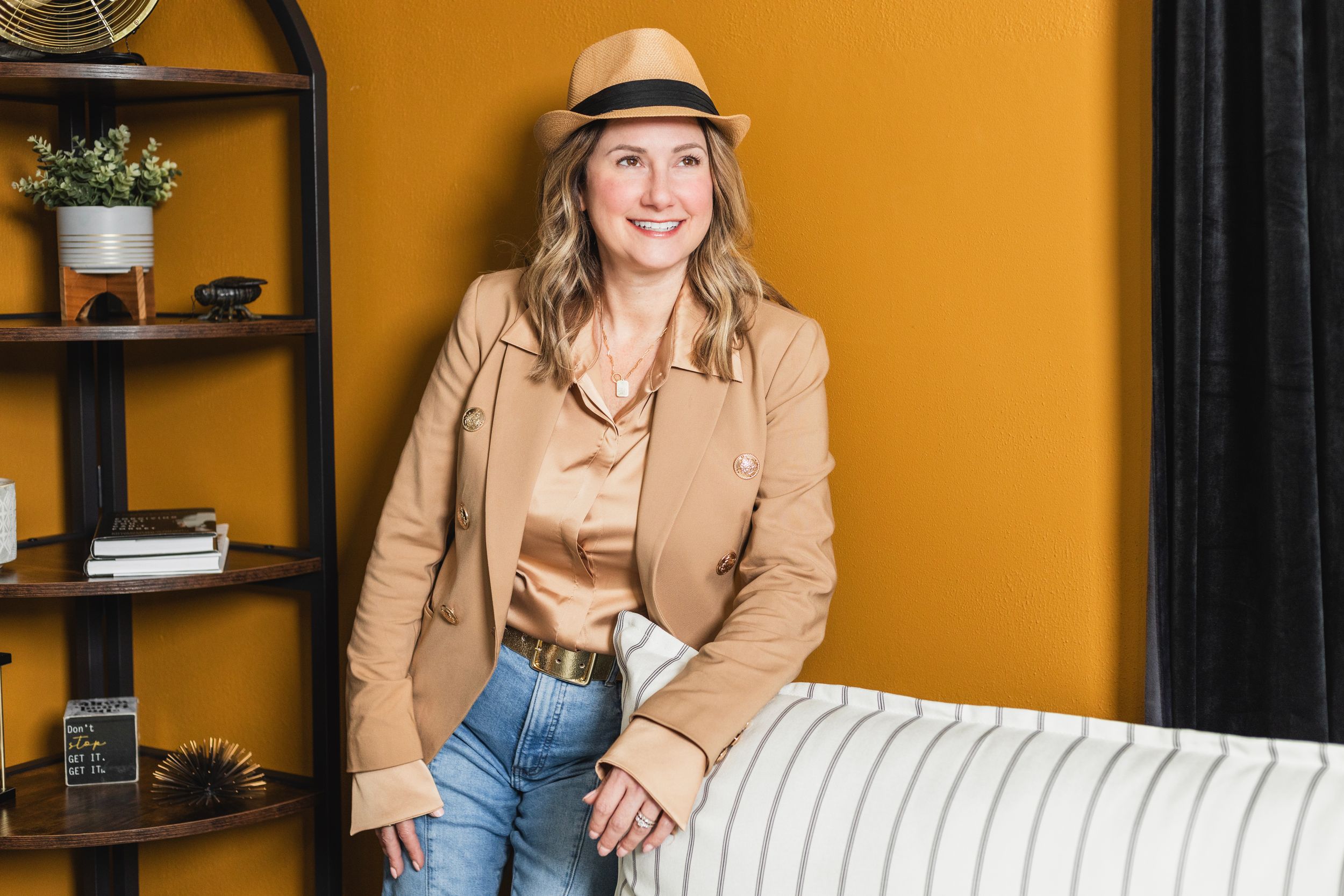 Professional wearing a tan blazer and hat poses casually against an orange wall in an office setting.
