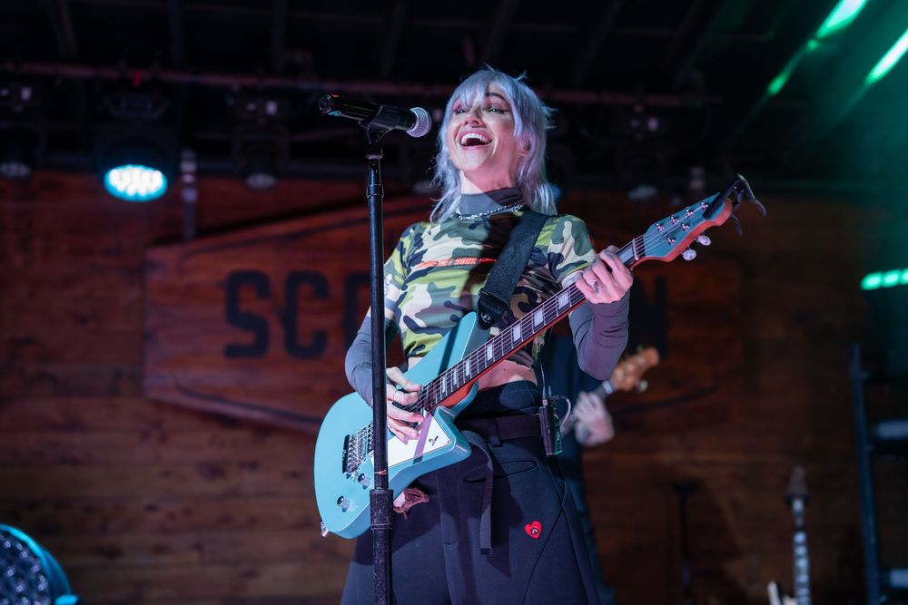 Blonde female guitarist in camo top playing teal electric guitar on stage, laughing joyfully.