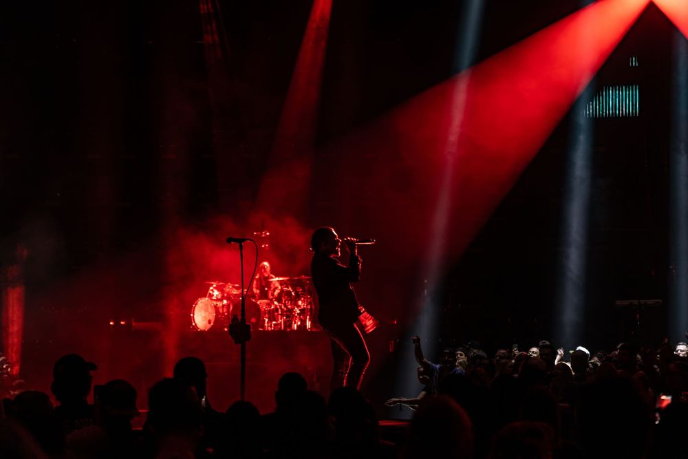 Rock band performing on a dramatic red-lit stage with spotlight beams and silhouetted crowd at a live concert.