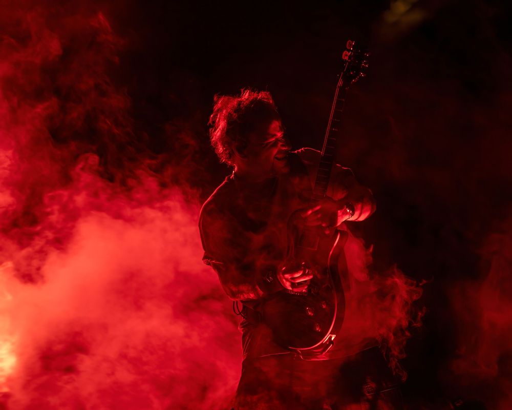 Guitarist performing on stage surrounded by dramatic red smoke and fog in a dark concert setting.