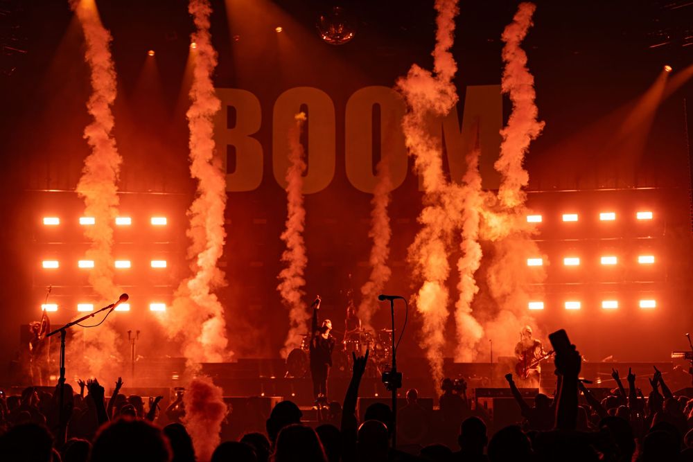 Rock concert stage with dramatic red pyrotechnics, smoke effects, and BOOM text display lighting up the arena.