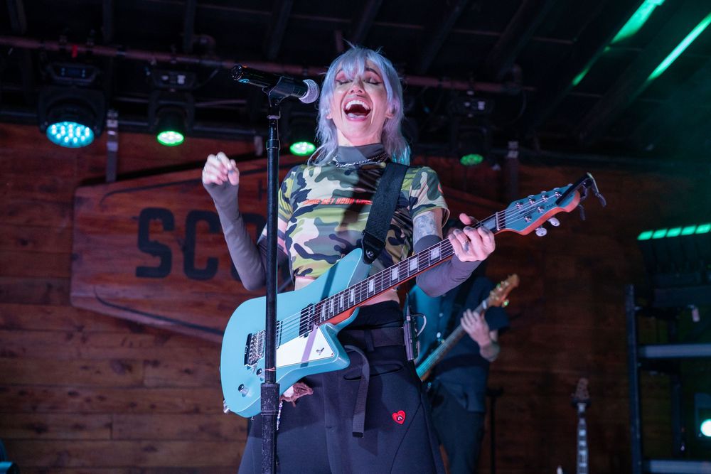 Energetic blonde female guitarist in camo top playing teal guitar on stage with arms raised.
