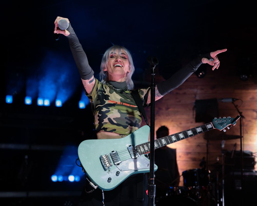 Female rock musician in camo top raising arm triumphantly while playing teal guitar on stage.