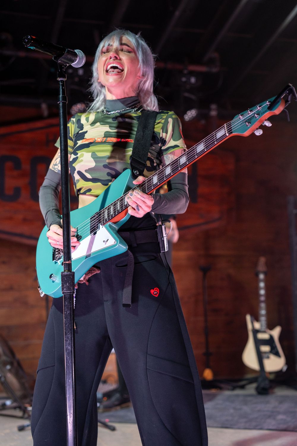Blonde female guitarist in camo top passionately playing teal electric guitar on stage.