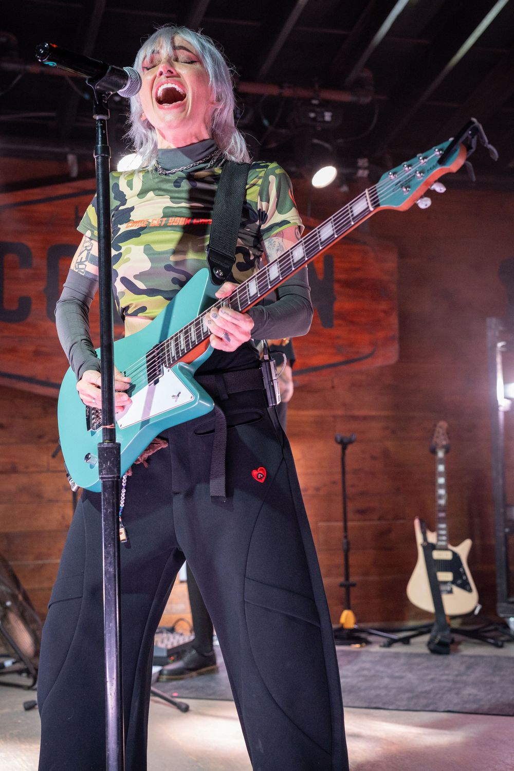 Female rock musician with blonde hair performing on stage with teal guitar in camo top.