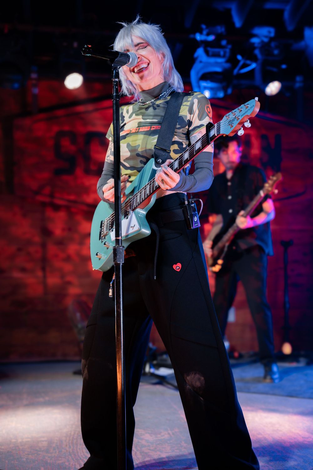 Female rock musician with blonde hair playing teal guitar on stage in camo top and black pants.