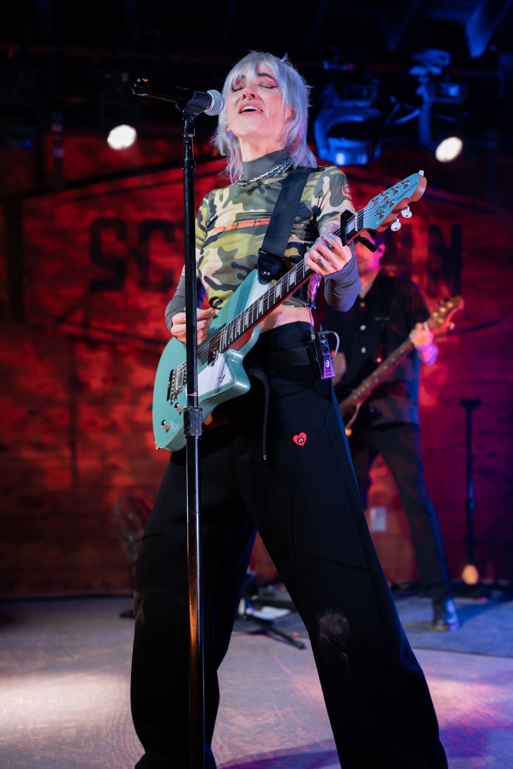 Female guitarist with blonde hair performing on stage, singing passionately with teal electric guitar.