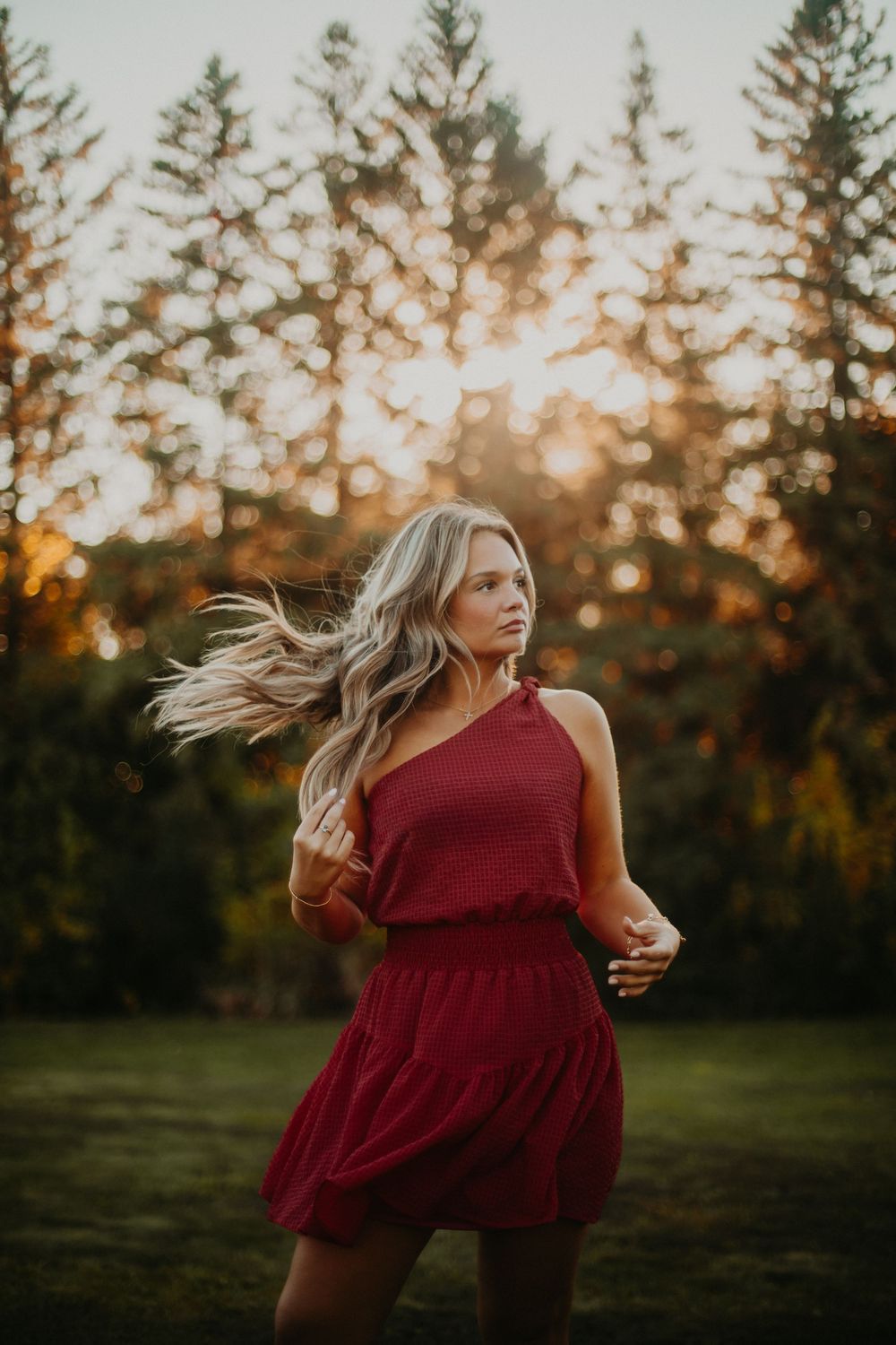 A person in a flowing burgundy dress twirls in a grassy field at sunset with evergreen trees in the background.
