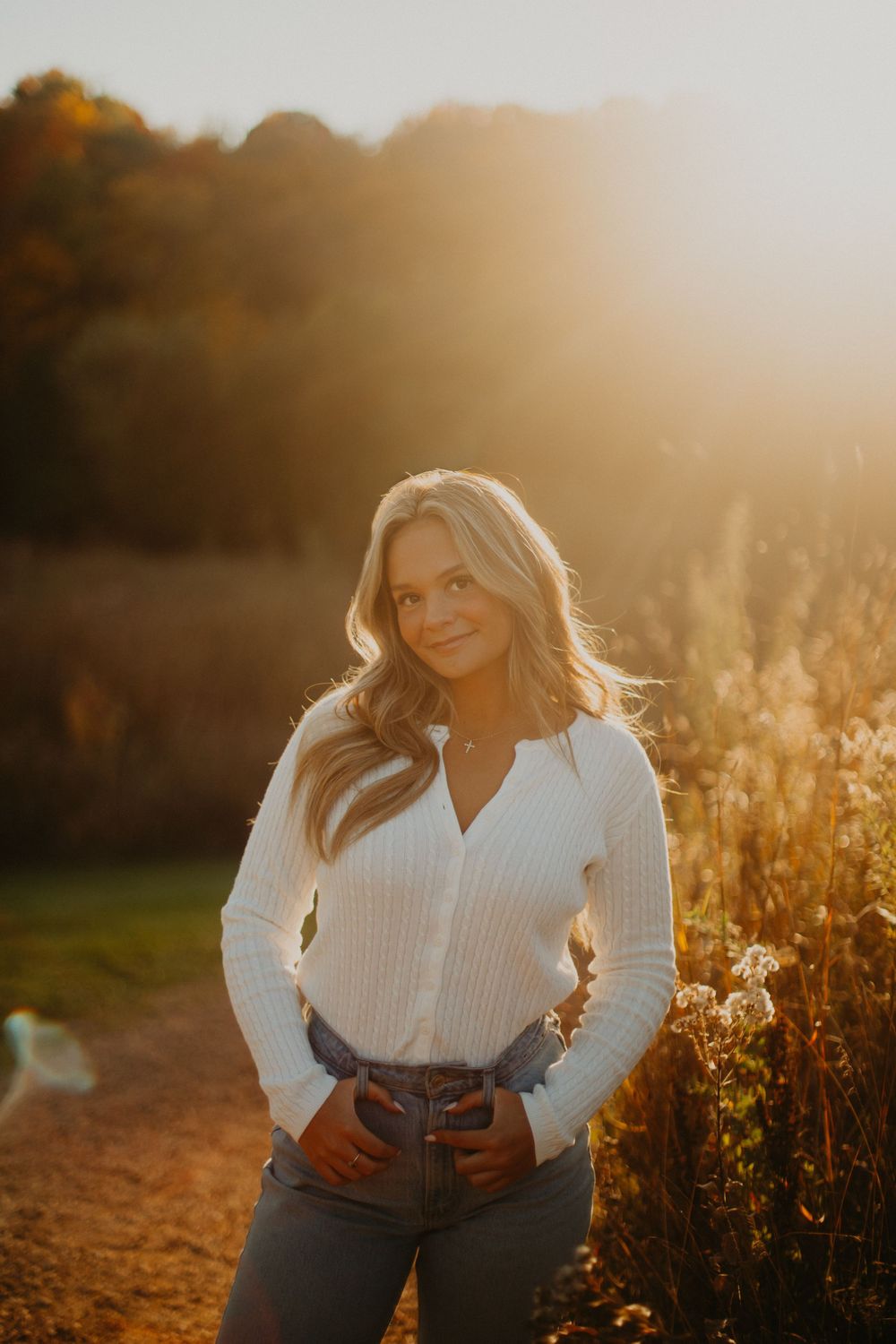 A person poses outdoors in a white sweater and jeans during golden hour with sunlit wildflowers in the background.