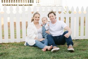 Happy pregnant couple sitting on grass in front of a white picket fence with their baby.