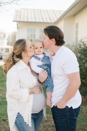Pregnant mother and father kissing their baby on the cheeks outdoors in front of a white house.