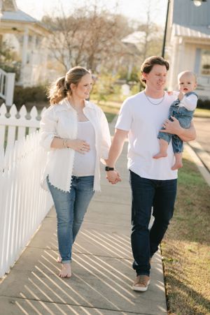 Smiling pregnant woman and man holding a baby walking hand in hand along a white picket fence.