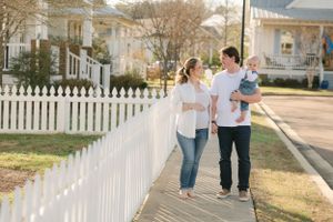 Young family with a baby strolling along a sidewalk beside a white picket fence in a neighborhood.