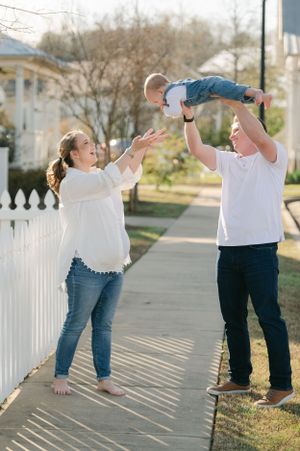 Father lifting baby in the air while pregnant mother reaches out on a sunny sidewalk.