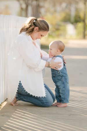 Pregnant mother kneeling beside white picket fence smiling at her standing baby in denim overalls.