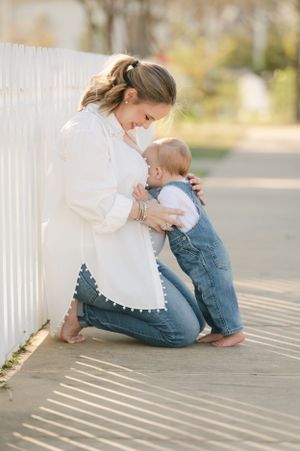 Pregnant mother sitting on a sidewalk cuddling her baby in denim overalls against a white fence.