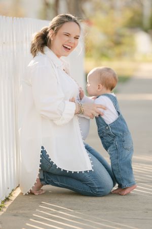 Laughing pregnant woman in white shirt and jeans kneeling by white fence with baby in denim overalls.