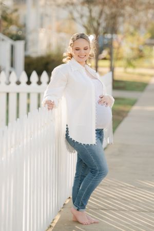Pregnant woman in white blouse and jeans leaning against white picket fence outdoors in sunlight.