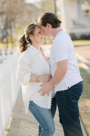 Couple embracing tenderly by white picket fence outdoors, man kissing pregnant woman on forehead.