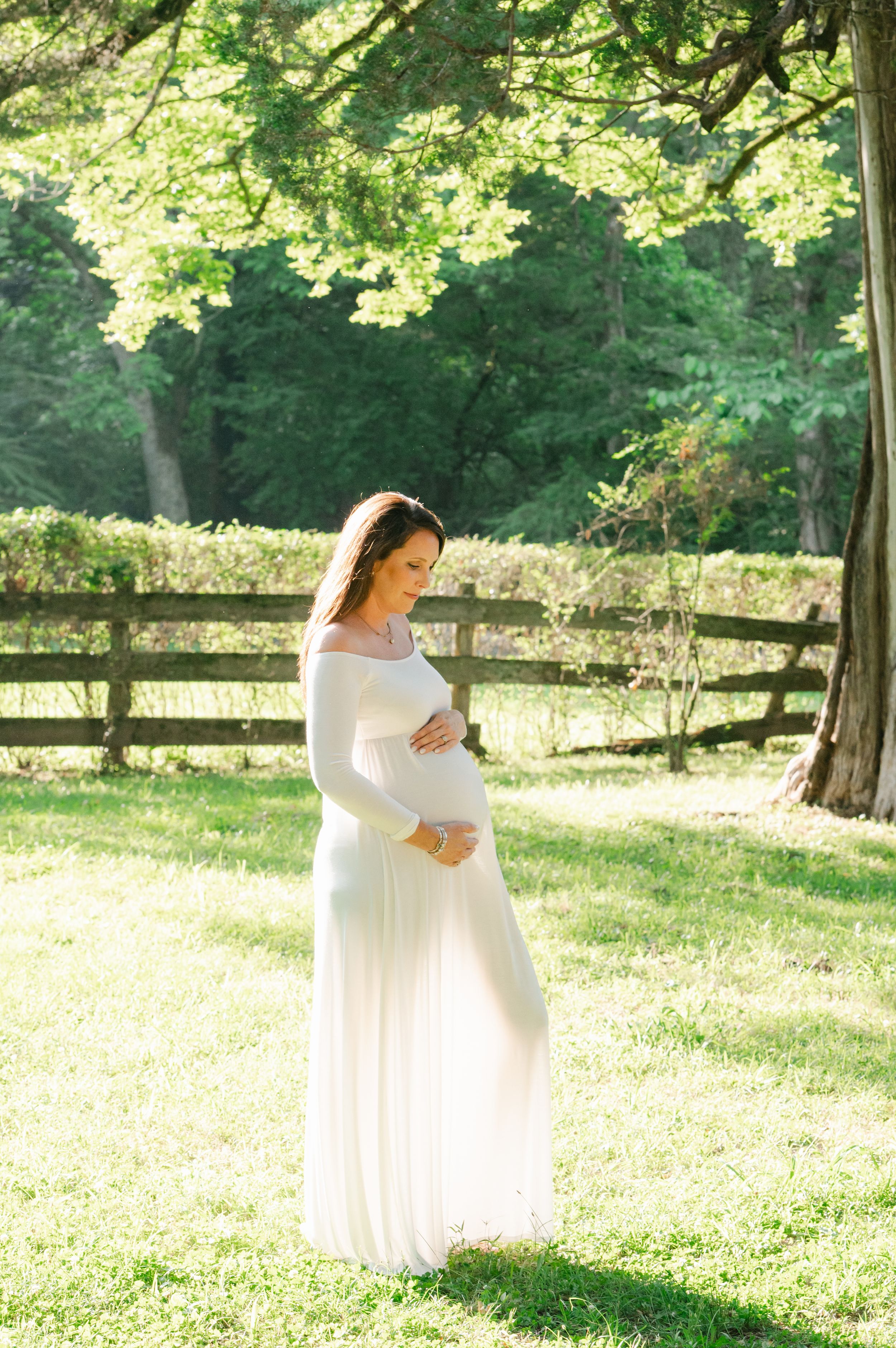 Pregnant woman in white off-shoulder gown cradling baby bump in sunlit green park setting.