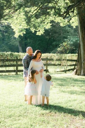 Family maternity photo with father, pregnant mother in white gown, and two young children outdoors.