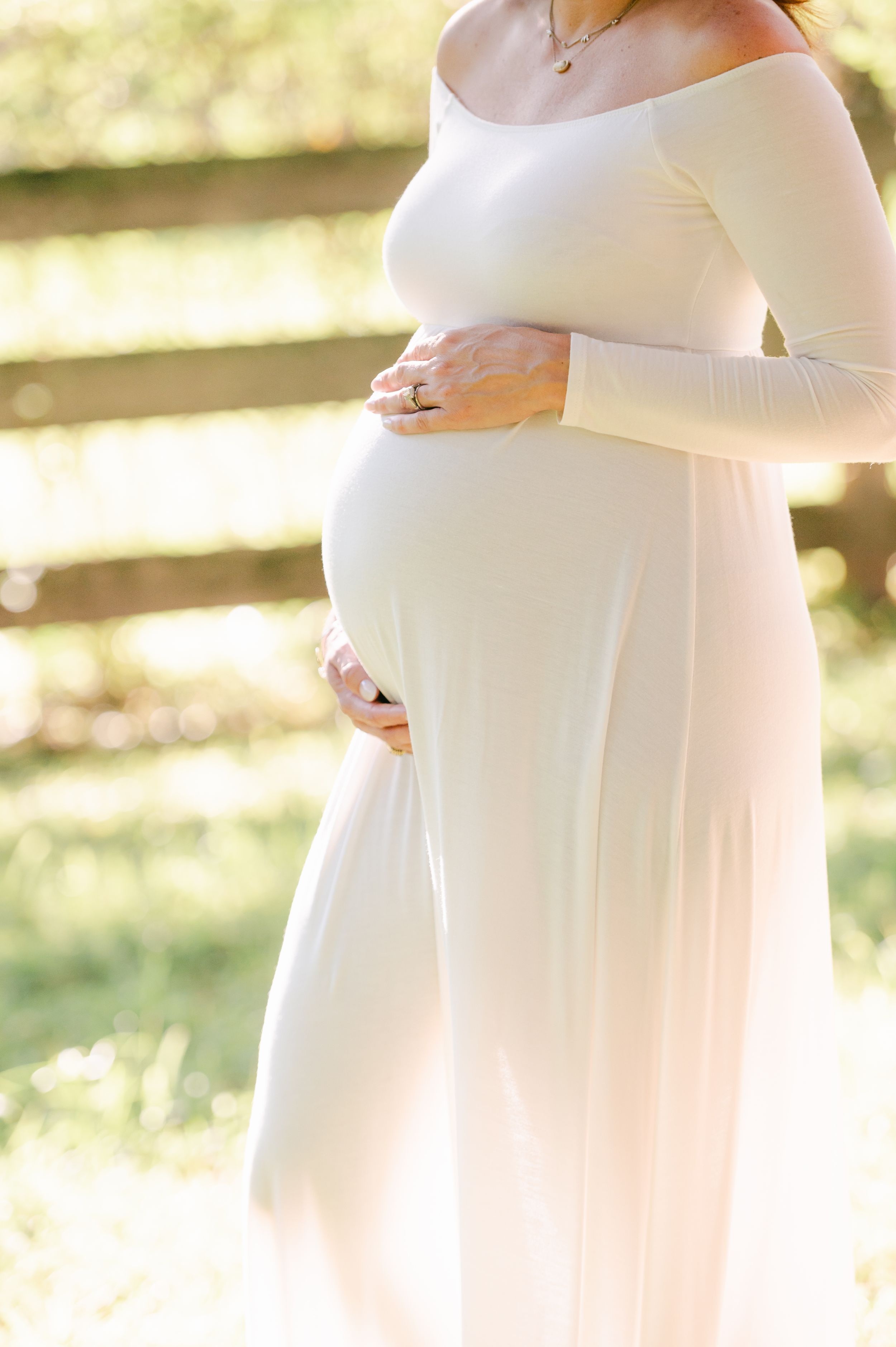 Close-up of pregnant woman in white off-shoulder dress gently holding baby bump outdoors.