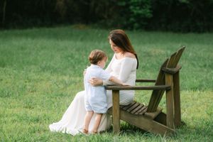 Pregnant woman in white gown sitting on wooden chair in grass with young child beside her.