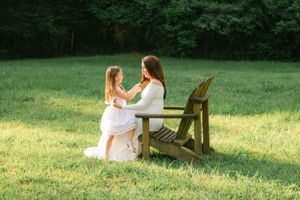 Pregnant woman in white dress sitting on wooden chair interacting with young girl in sunny meadow.