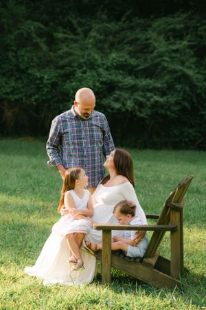 Family of four with pregnant mother seated on wooden chair in lush green outdoor setting.