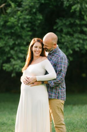 Smiling pregnant woman in white gown embraced from behind by bald man in plaid shirt outdoors.