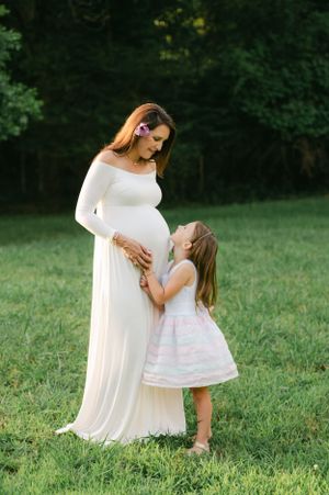 Pregnant woman in white off-shoulder gown with daughter in pink dress sharing tender moment outdoors.