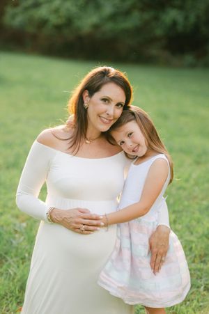 Pregnant woman in white maternity gown smiling with young daughter hugging her in sunny green field.