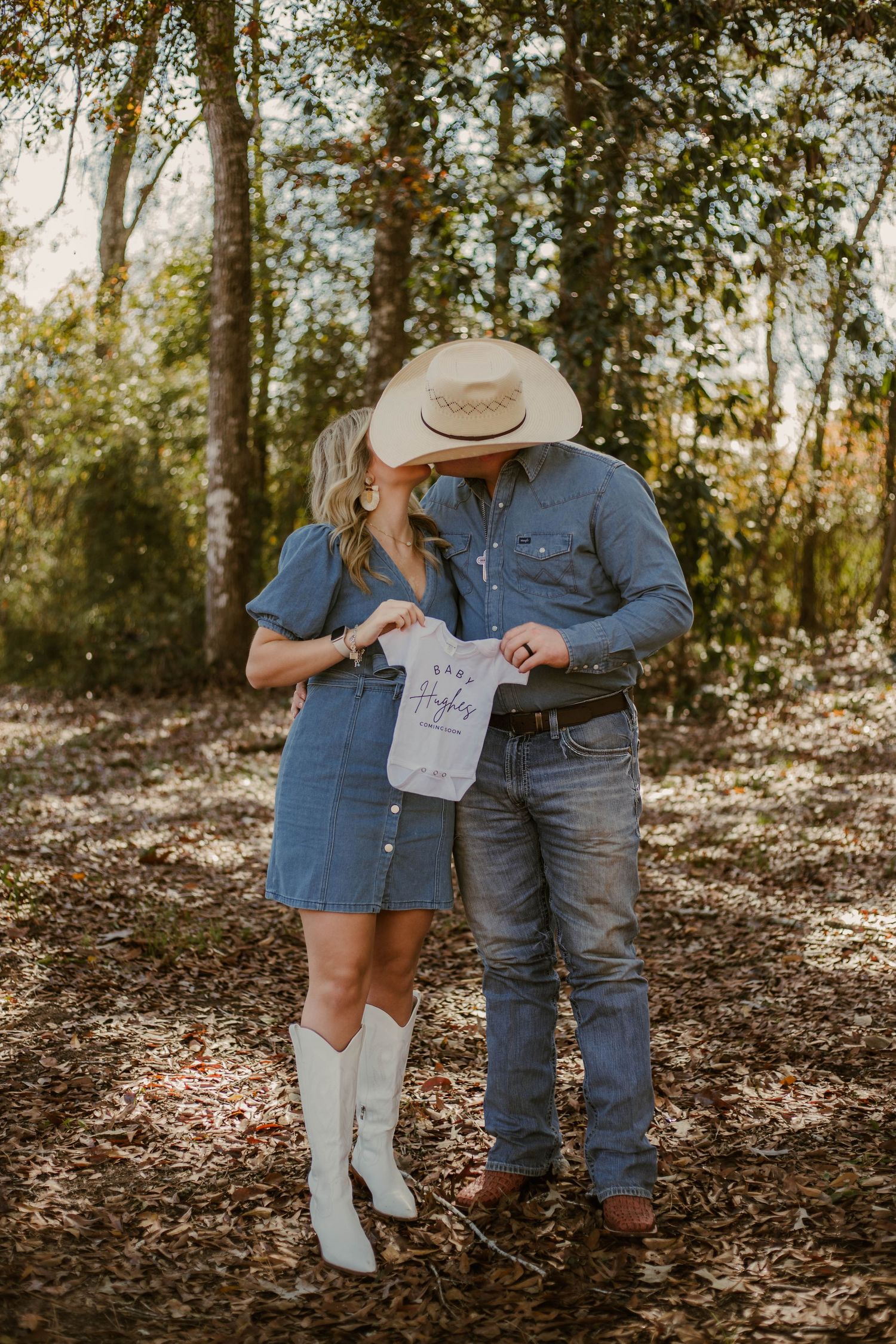 Couple in denim outfits share intimate moment while holding baby announcement sign in wooded setting.