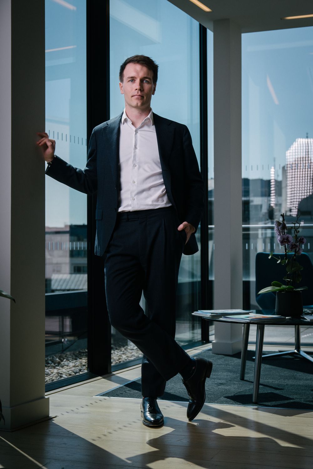 Modern office portrait with business professional in dark suit against city window backdrop.