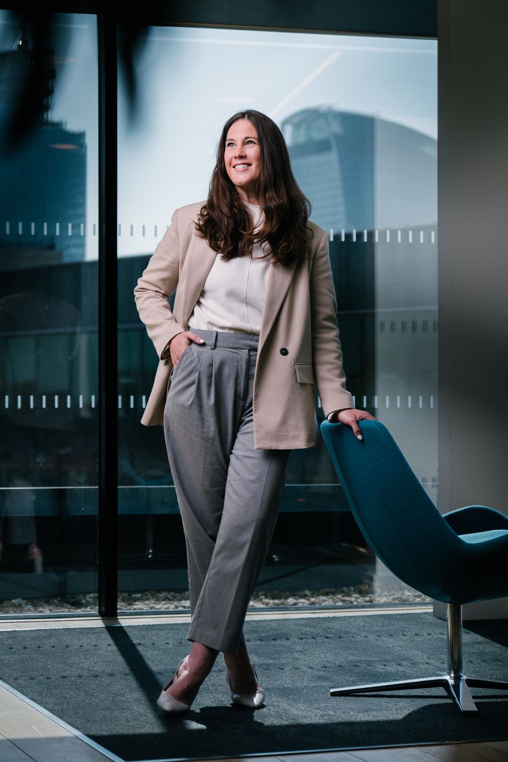 Professional wearing beige blazer and gray pants poses confidently in modern office setting.