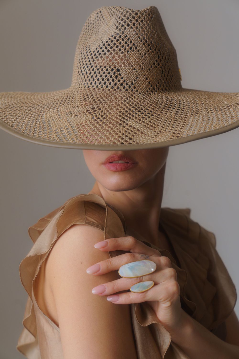 Artistic portrait featuring a wide-brim straw hat and statement ring against neutral backdrop.