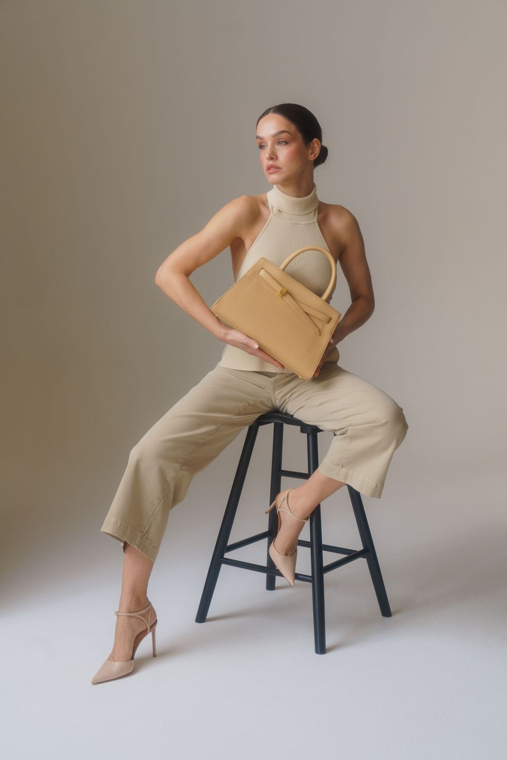 Minimalist fashion photo with model in beige outfit holding tan leather handbag while posed on stool.