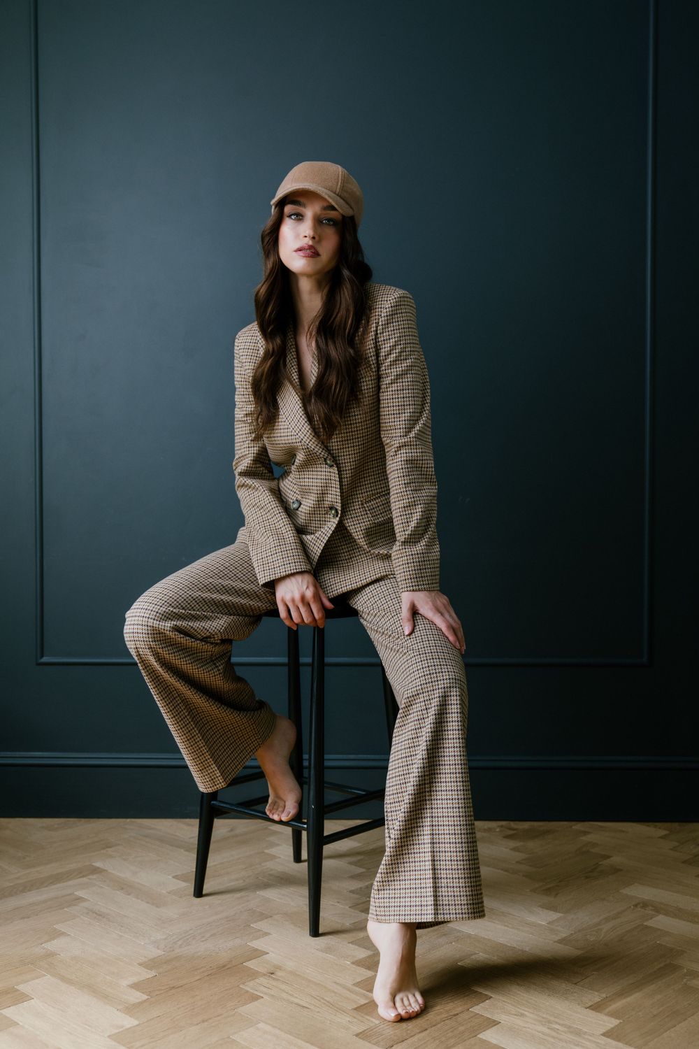 A fashion model poses on a stool wearing a plaid pantsuit and beige cap against a dark backdrop.
