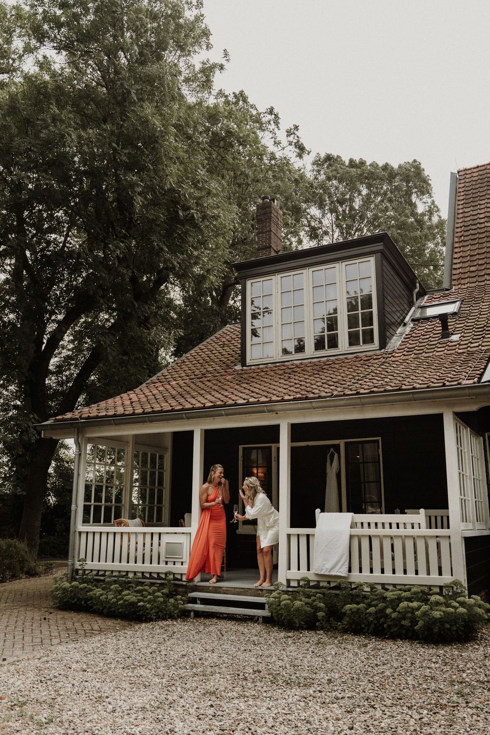 Charming cottage with white porch railing and dormer window sits among trees with gravel driveway.