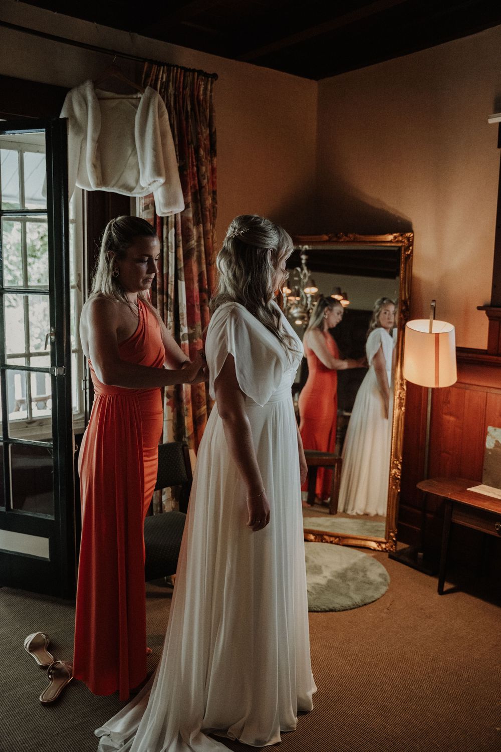 A bridal party gets ready in a cozy room with warm lighting and a full-length mirror reflecting the elegant dresses.