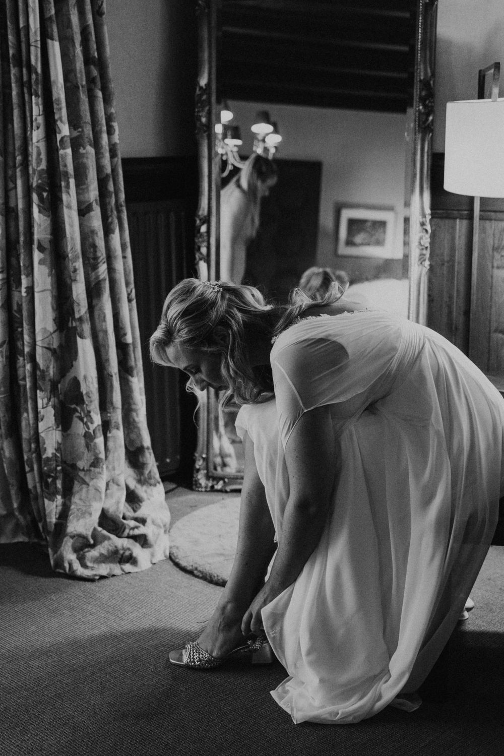 A person in a flowing white dress bends down to adjust their shoe near floral curtains in a dimly lit room.